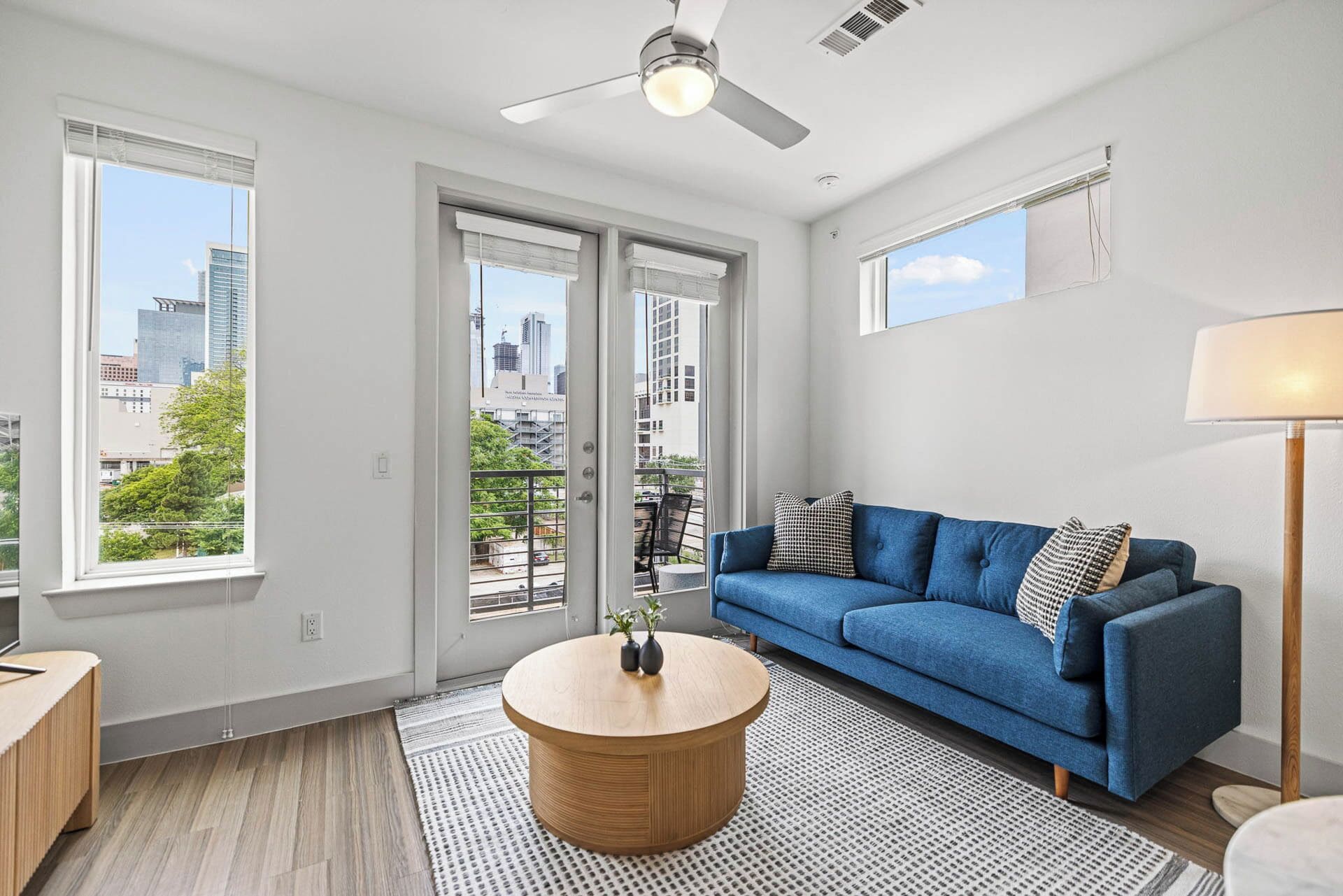 A modern living room with a blue sofa, a round wooden coffee table, and a city view through large windows.