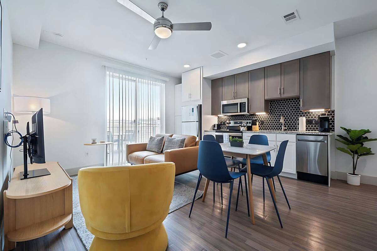 A modern apartment with a living room, dining area, and kitchen. There is a sofa, chairs, a TV, and a ceiling fan.