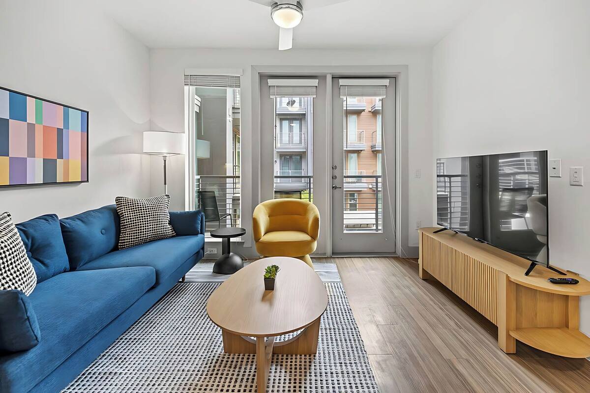 A modern living room with a blue sofa, a yellow armchair, a wooden coffee table, and a TV on a wooden stand.
