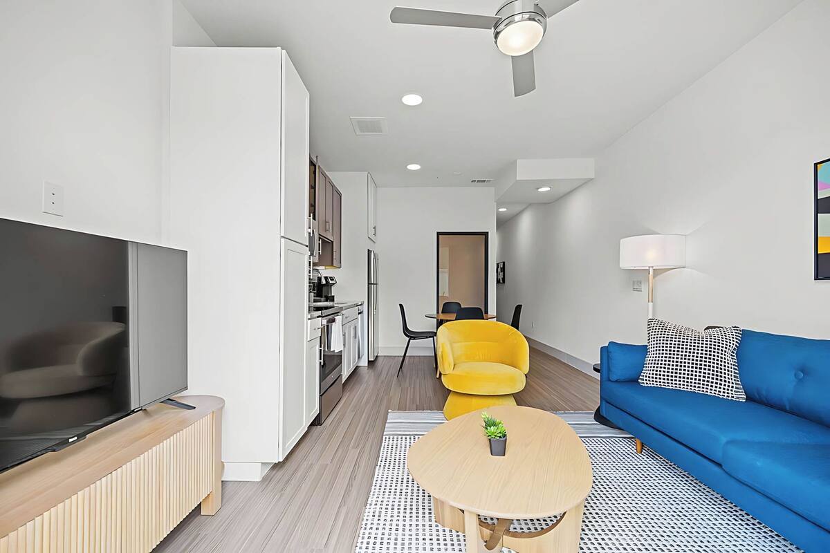 A modern living room with a blue sofa, a yellow armchair, a small wooden table with a plant, and a TV on a wooden stand.