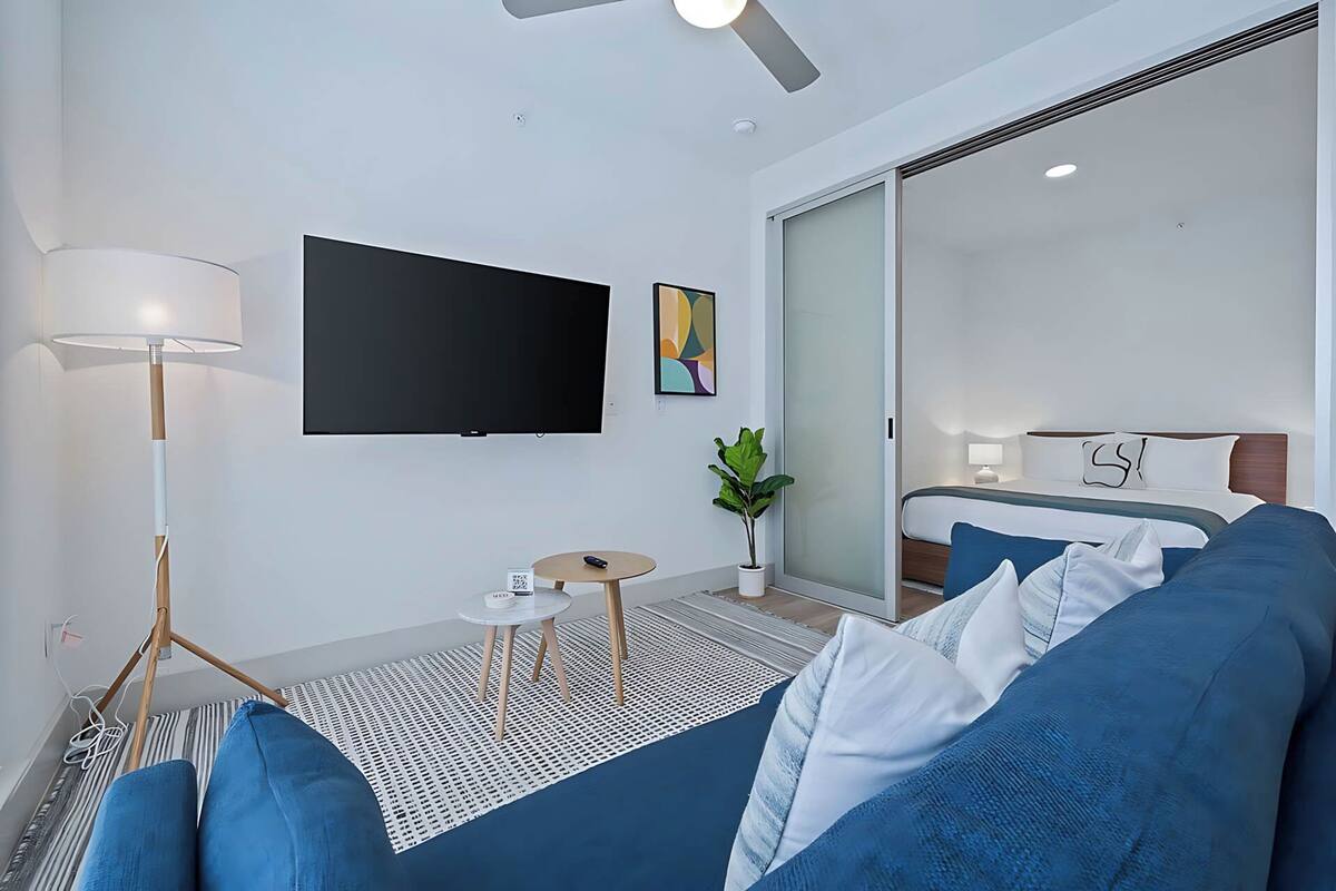 A modern hotel room with a large bed, a sofa, a flat-screen TV, a floor lamp, and a small table.
