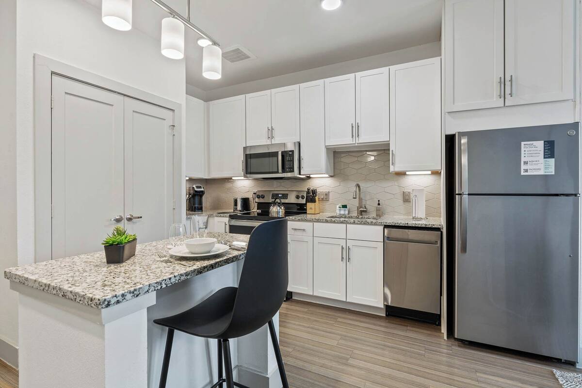 A modern kitchen with white cabinets, stainless steel appliances, a granite countertop, and a black bar stool.