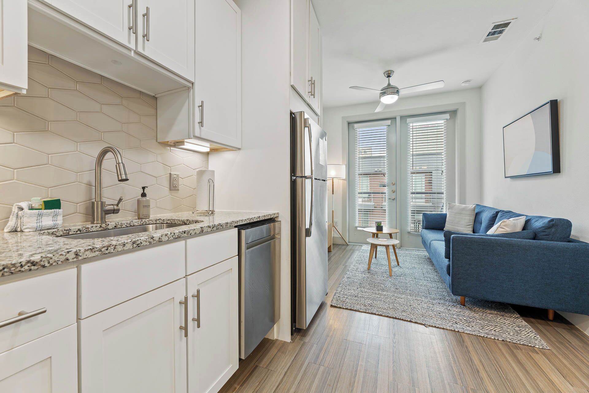 A modern kitchen with white cabinets, a granite countertop, and stainless steel appliances. There is a blue sofa and a small table in the living area, with a patterned rug on the floor.