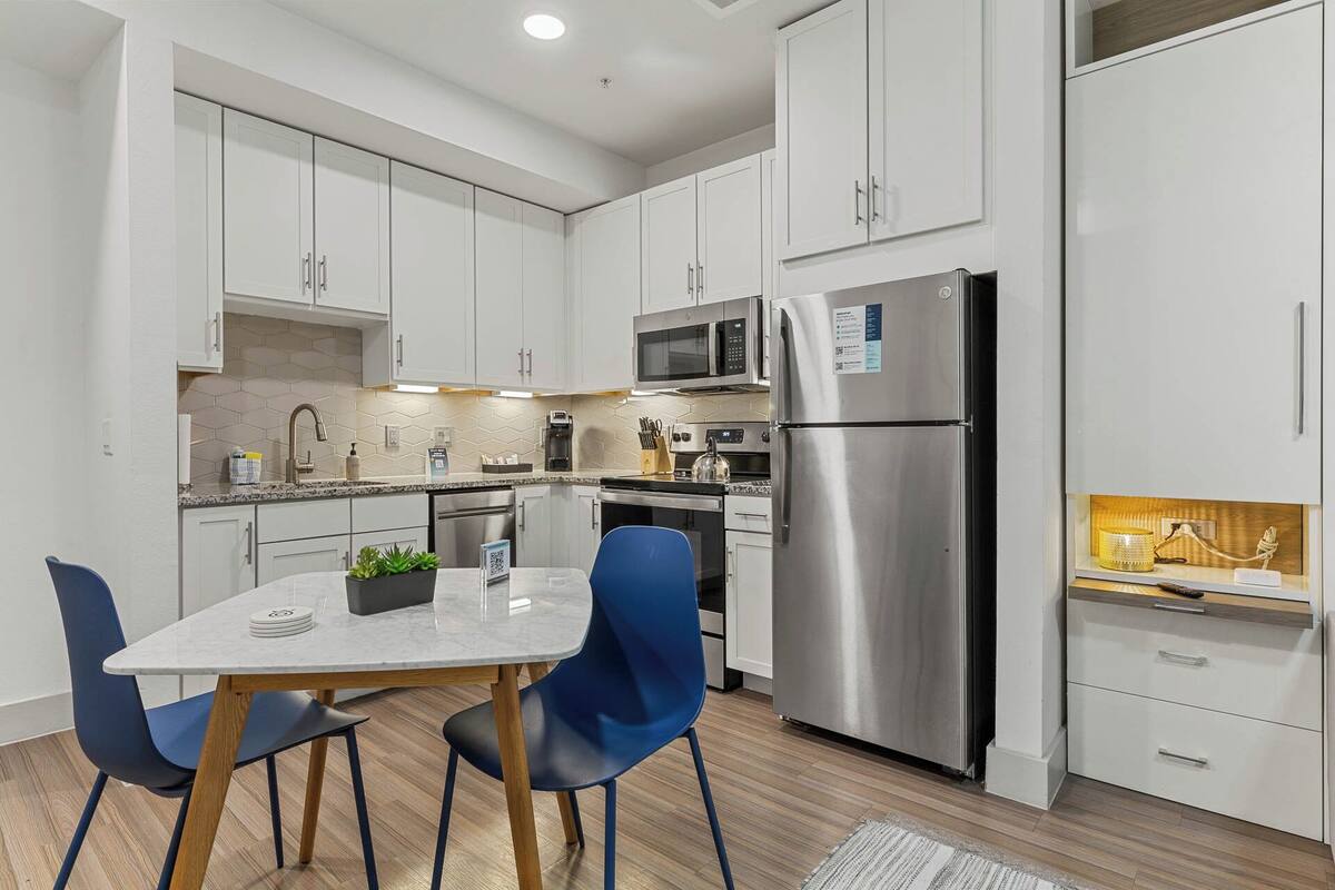 A modern kitchen with white cabinets, stainless steel appliances, a marble countertop, and blue chairs.