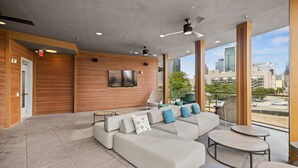 A modern outdoor lounge area with a large sectional sofa, a flat-screen TV mounted on the wall, and a view of the city skyline. - Placemakr Austin Downtown, Apartments by Hilton (Austin)