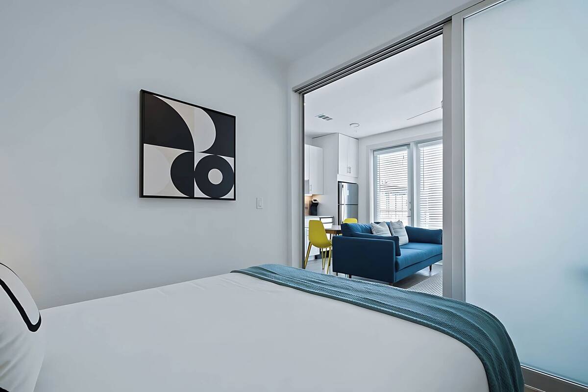 A modern hotel room with a bed, a large abstract painting, a view of the living area, and a sliding door leading to the balcony.