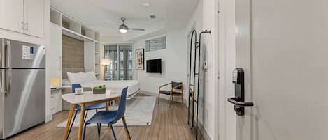 Una habitación de hotel moderna con kitchenette, una zona de comedor con mesa y sillas, cama, televisión y una puerta corredera con cerradura electrónica.