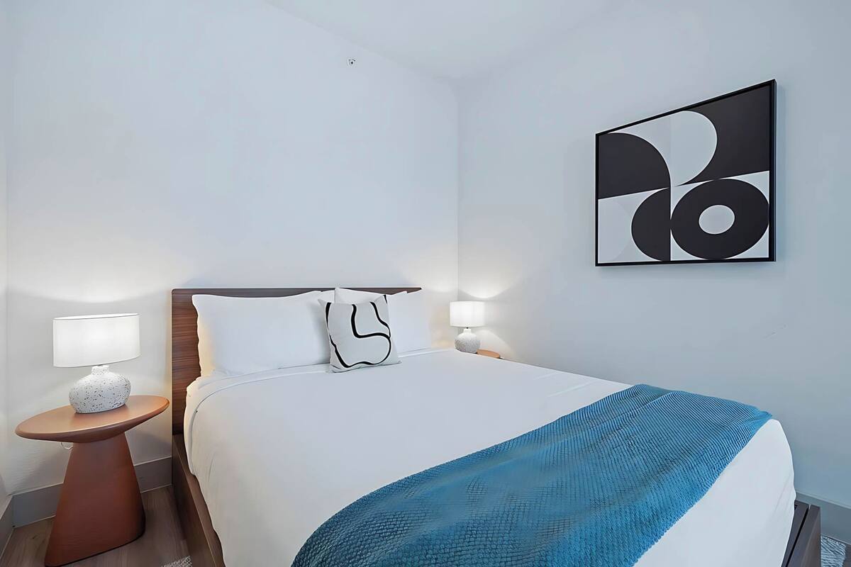 A hotel room with a bed, two bedside tables with lamps, a wall-mounted abstract artwork, and a blue textured blanket.