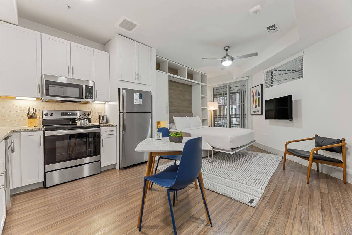 A compact hotel room with a kitchenette, a bed, a TV, and a ceiling fan.