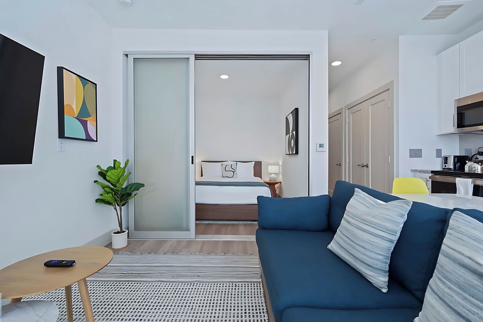 A modern, compact living space with a sofa, a small table, a potted plant, and a sliding door leading to a bedroom.