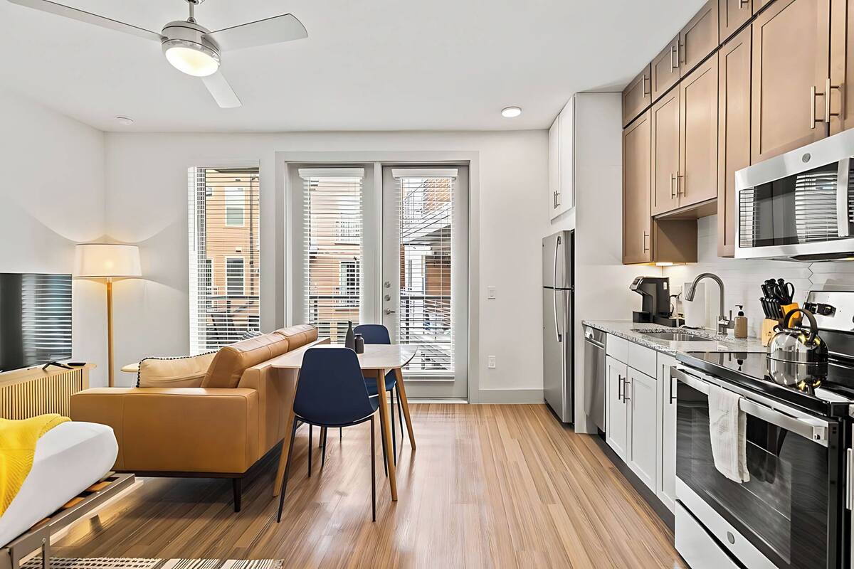 A modern kitchen with stainless steel appliances, a dining area with a sofa and chairs, and a large window with blinds.