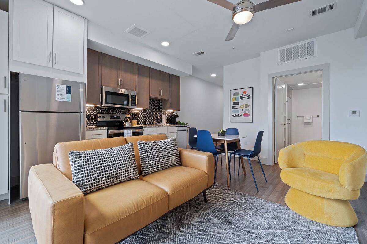 A modern kitchen with a microwave, stove, and refrigerator. There is a dining area with a table and chairs, and a yellow armchair.