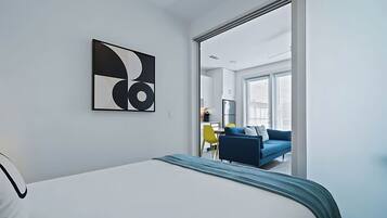 A modern hotel room with a bed, a large abstract artwork, a view of the dining area, and a sliding door leading to a balcony.