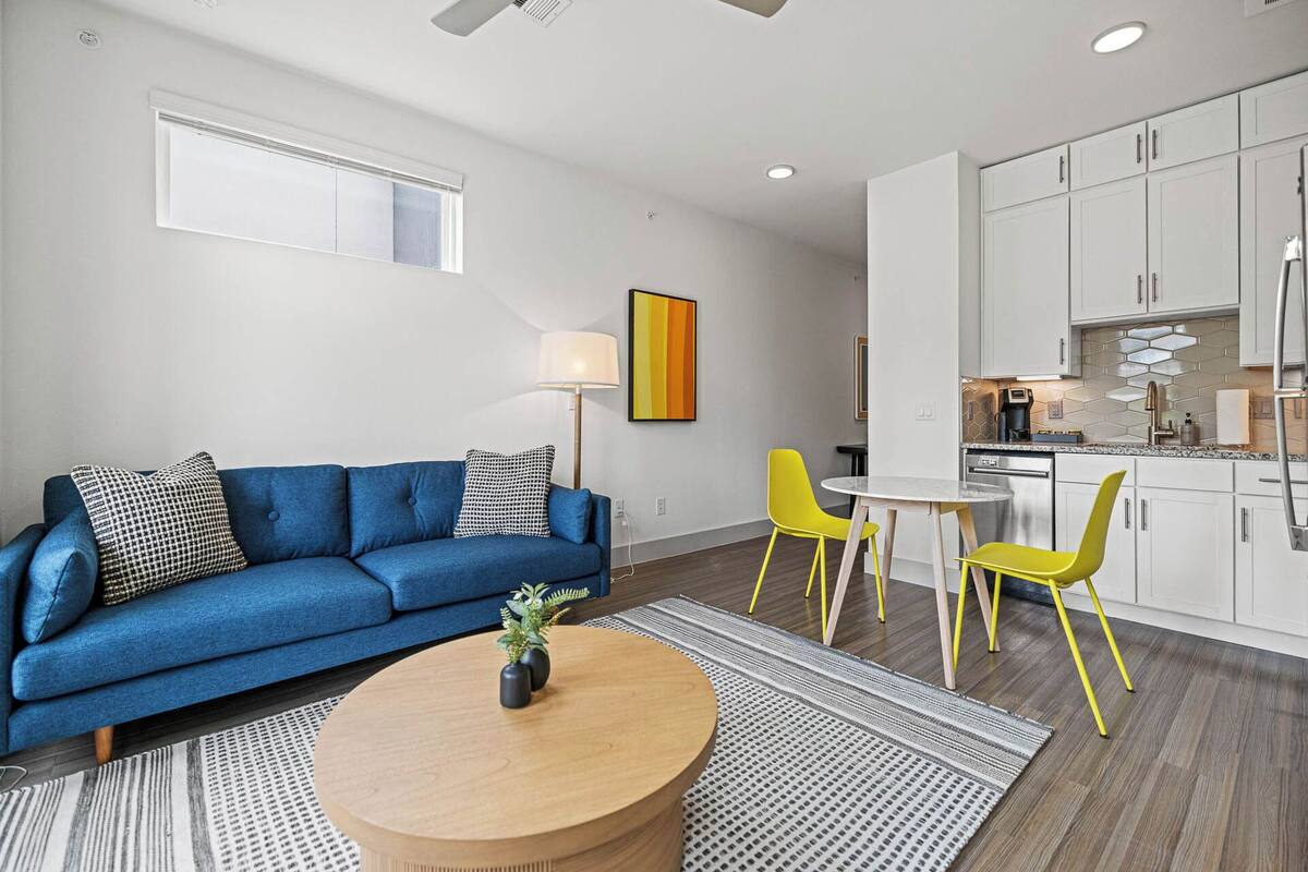 A modern living room with a blue sofa, yellow chairs, a round wooden coffee table, and a kitchen area with white cabinets and a stainless steel refrigerator.