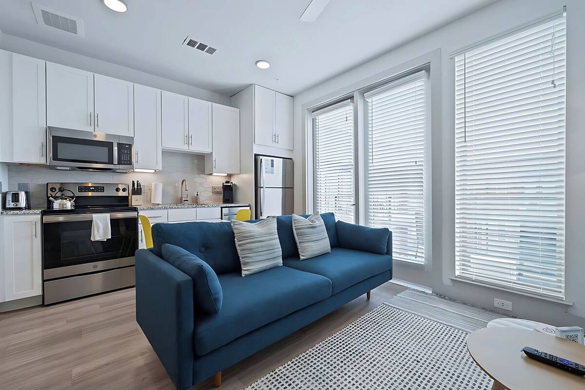 A modern kitchen with white cabinets, a microwave, and a stove. There is a blue sofa with pillows and a small table with a remote control.