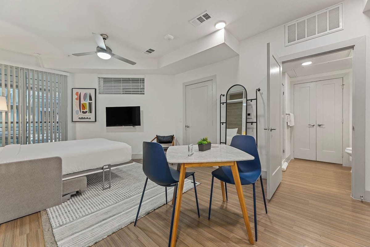 A modern, well-lit room with a bed, a TV, a dining table with chairs, a ceiling fan, and a mirror.