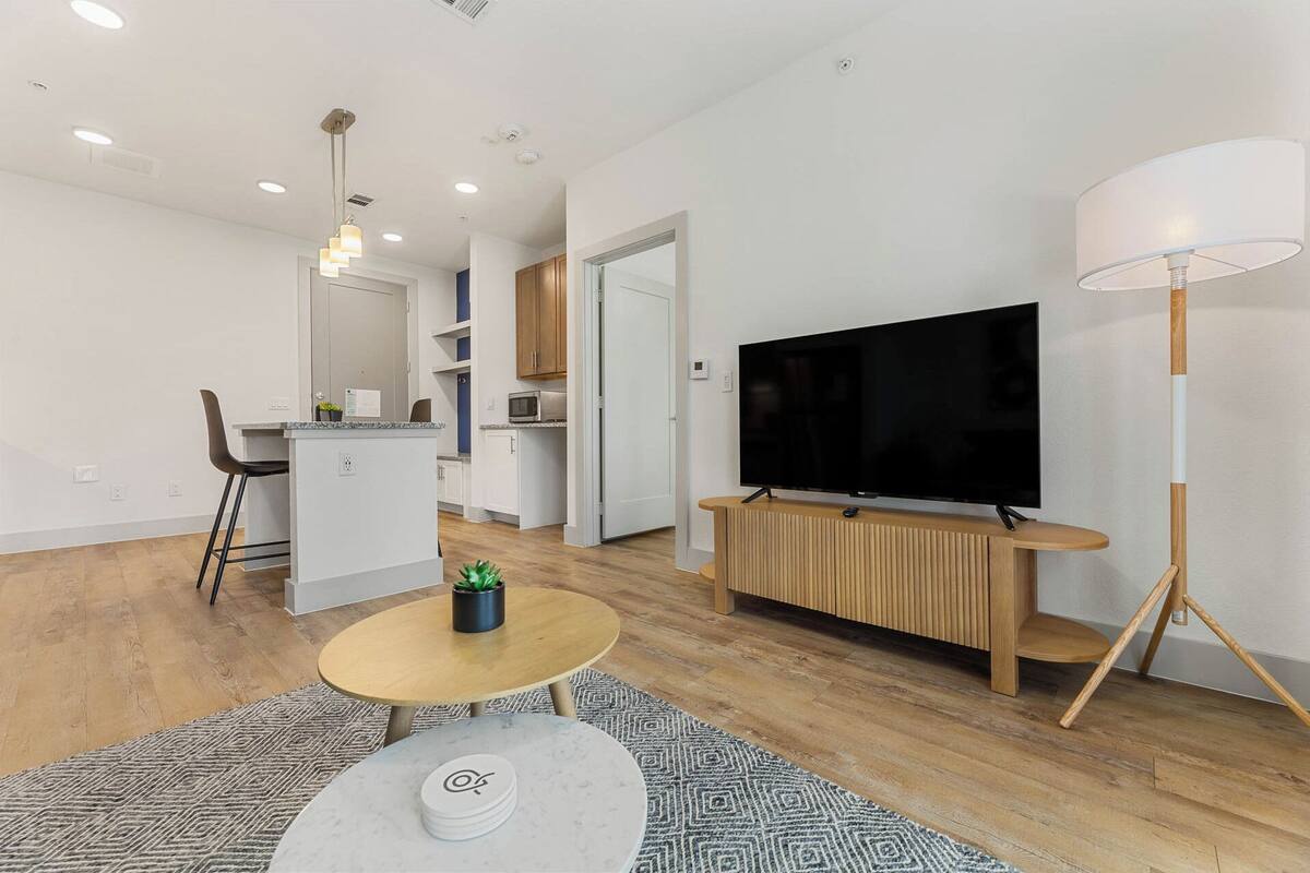 A modern living room with a flat-screen TV on a wooden stand, a dining area with a small kitchen, and a large floor lamp.