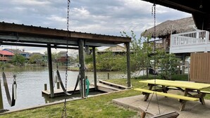 Ein Holzschaukelstuhl, ein Picknicktisch und ein überdachter Bereich mit Blick auf ein Gewässer und Gebäude im Hintergrund.