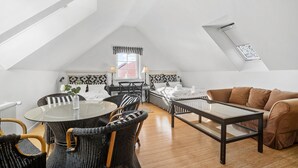Ein geräumiges Schlafzimmer im Loft-Stil mit schrägen Wänden, einem großen Dachfenster, einem Bett mit schwarz-weiß gestreifter Bettwäsche, einem Glastisch, Korbstühlen und einem braunen Ledersofa.