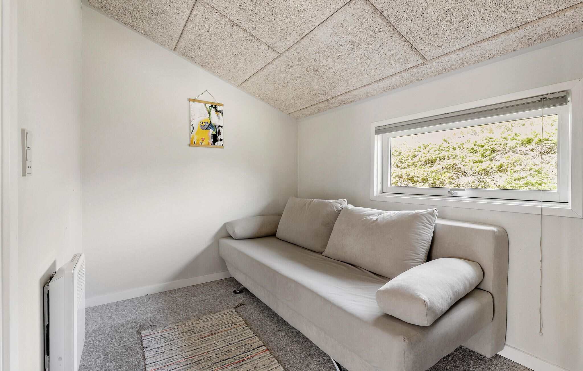 A room with a beige sofa, a hanging abstract painting, a radiator, a window with a view of greenery, and a textured ceiling.