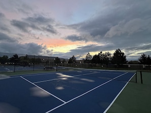 Ulkotenniskenttä, jossa on verkko, ja jota ympäröivät puut ja pilvinen taivas.