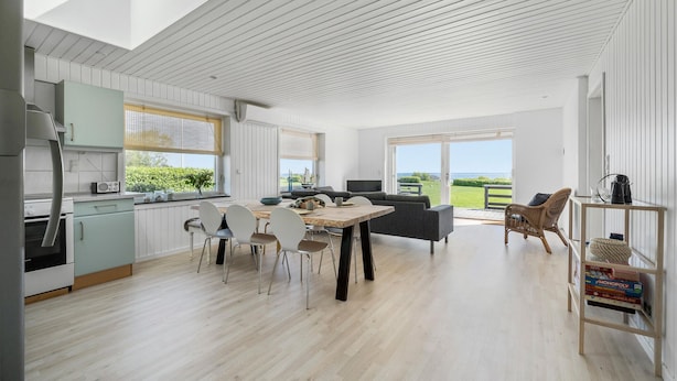 A modern kitchen with white cabinetry, a dining area with a wooden table and chairs, and a view of the sea through large windows. - Nice home in Hesselager with WiFi (Hesselager)