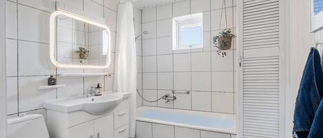 Een moderne badkamer met een witte wastafel, een badkamermeubel en een badkuip. Er is een raam met uitzicht, een hangplant en een handdoekhouder.