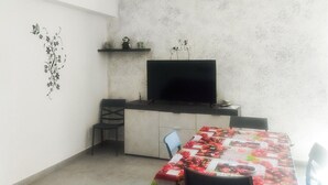 Un espacio habitable pequeño y funcional con kitchenette, un comedor con mantel floral y un televisor montado en la pared.