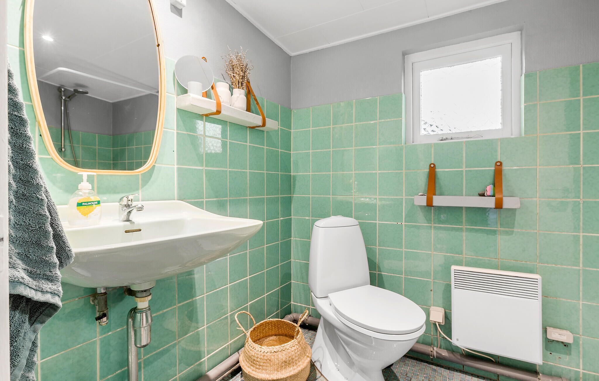 Een badkamer met een witte wastafel, toilet en spiegel. Er zijn planken met toiletartikelen en een raam boven het toilet.