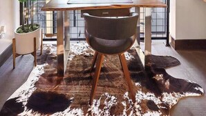 Un loft moderne avec un plafond en pente, un coin repas avec une table et des chaises en bois, une grande fenêtre donnant sur la forêt et un tapis en peau de vache sur le sol.