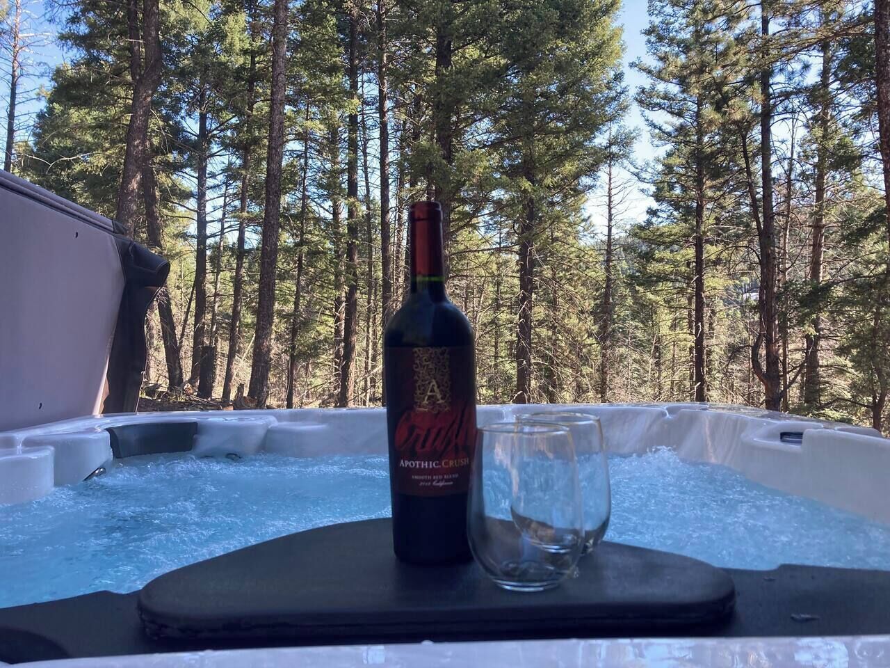 Un jacuzzi con una botella de vino y una copa sobre una mesa, rodeado de un bosque.