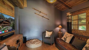 A cozy living room with a wooden ceiling, a television mounted on a wooden cabinet, a leather sofa, a small round ottoman, and a wooden side table with a lamp. - La Grange au Merle - mountain break for 12 with spa - OVO Network (Châtel)