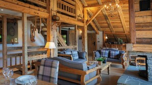 A rustic wooden interior with a high ceiling, exposed beams, and a stone fireplace. There are two sofas with plaid cushions and a dining table with wine glasses and a plant. - La Grange au Merle - mountain break for 12 with spa - OVO Network (Châtel)