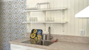 A kitchen with a sink, a countertop, and a backsplash with a floral pattern. There are two shelves above the sink, one holding glasses and the other holding a dish rack. - Awesome home in Västervik with kitchen (Västervik)