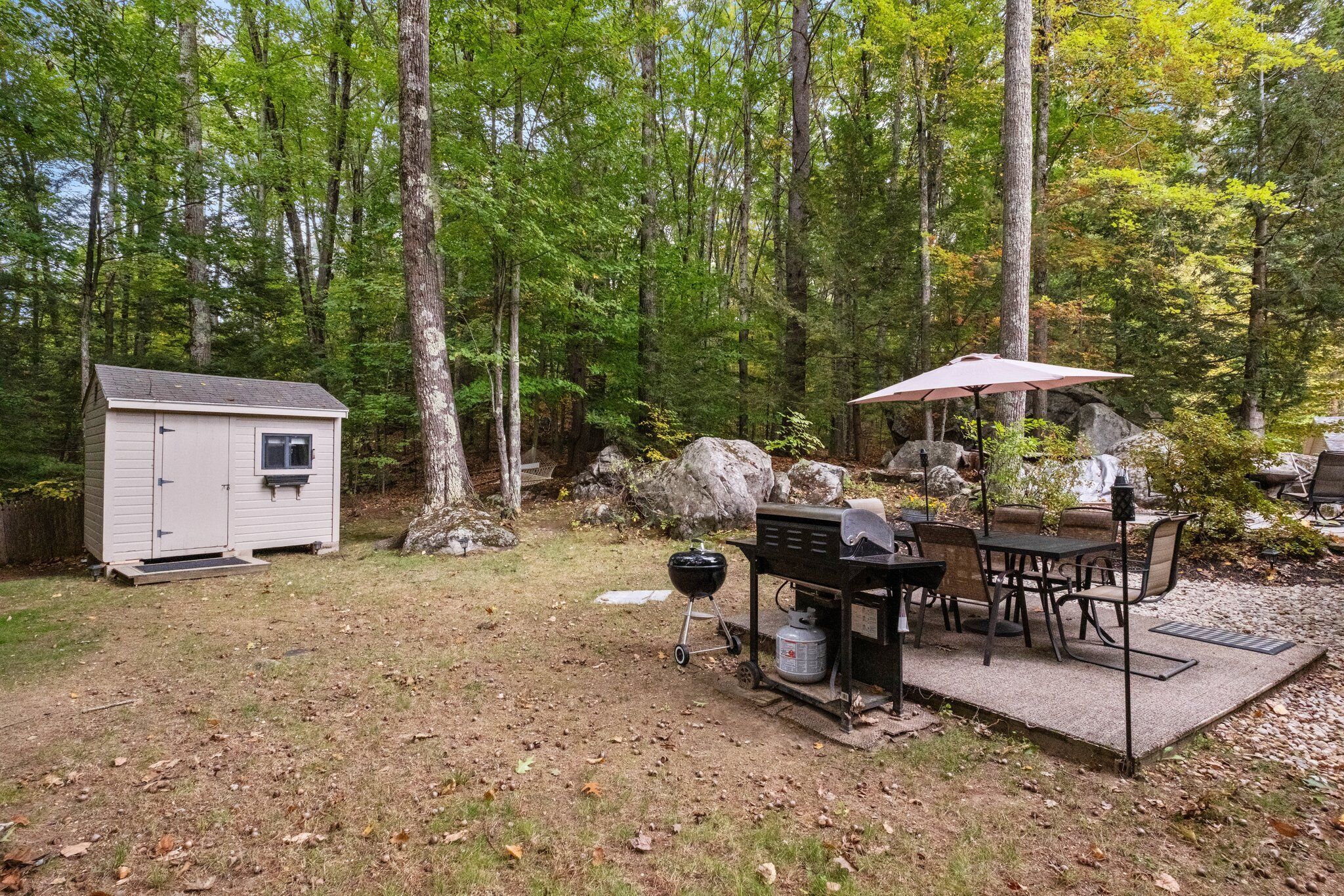 Un jardin avec un barbecue, une table à manger et des chaises sous un parasol, le tout entouré d’arbres et d’un abri de jardin.