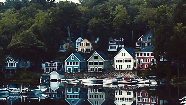 Un pueblo frente al agua con casas reflejadas en aguas tranquilas, rodeado de árboles frondosos.