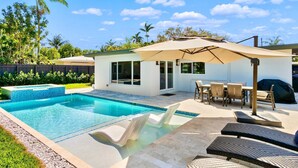 A backyard with a swimming pool, lounge chairs, a barbecue area, and a small patio with a table and chairs.