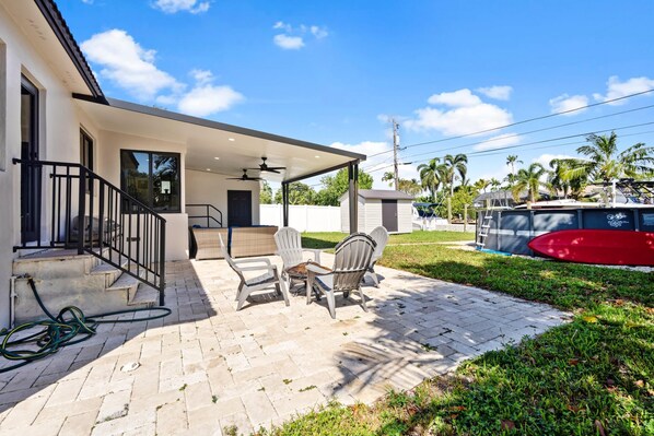 A modern house with a covered patio, outdoor seating, and a red boat in the background. - Stunning 3 Bd Pool House w Enchanting Water Views (Fort Lauderdale)