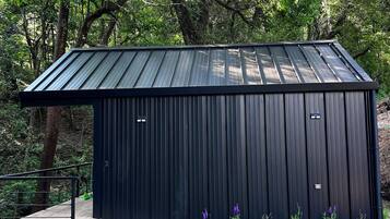 A black metal shed with a sloped roof, surrounded by a grassy area with small purple flowers and a gravel path leading to it.