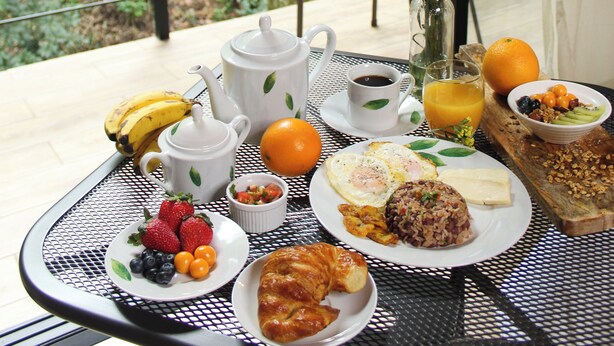 Une table de petit-déjeuner, dressée sur un balcon offrant une vue sur la forêt, est garnie d’une variété d’aliments et agrémentée d’un vase de fleurs.