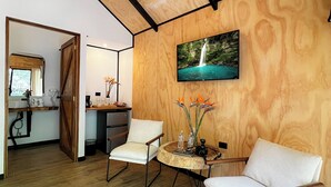 Un salon moderne avec un plafond en bois, un téléviseur fixé au mur, deux chaises blanches, une table ronde en bois sur laquelle se trouvent un appareil photo et des objets décoratifs, et une cuisine en arrière-plan.