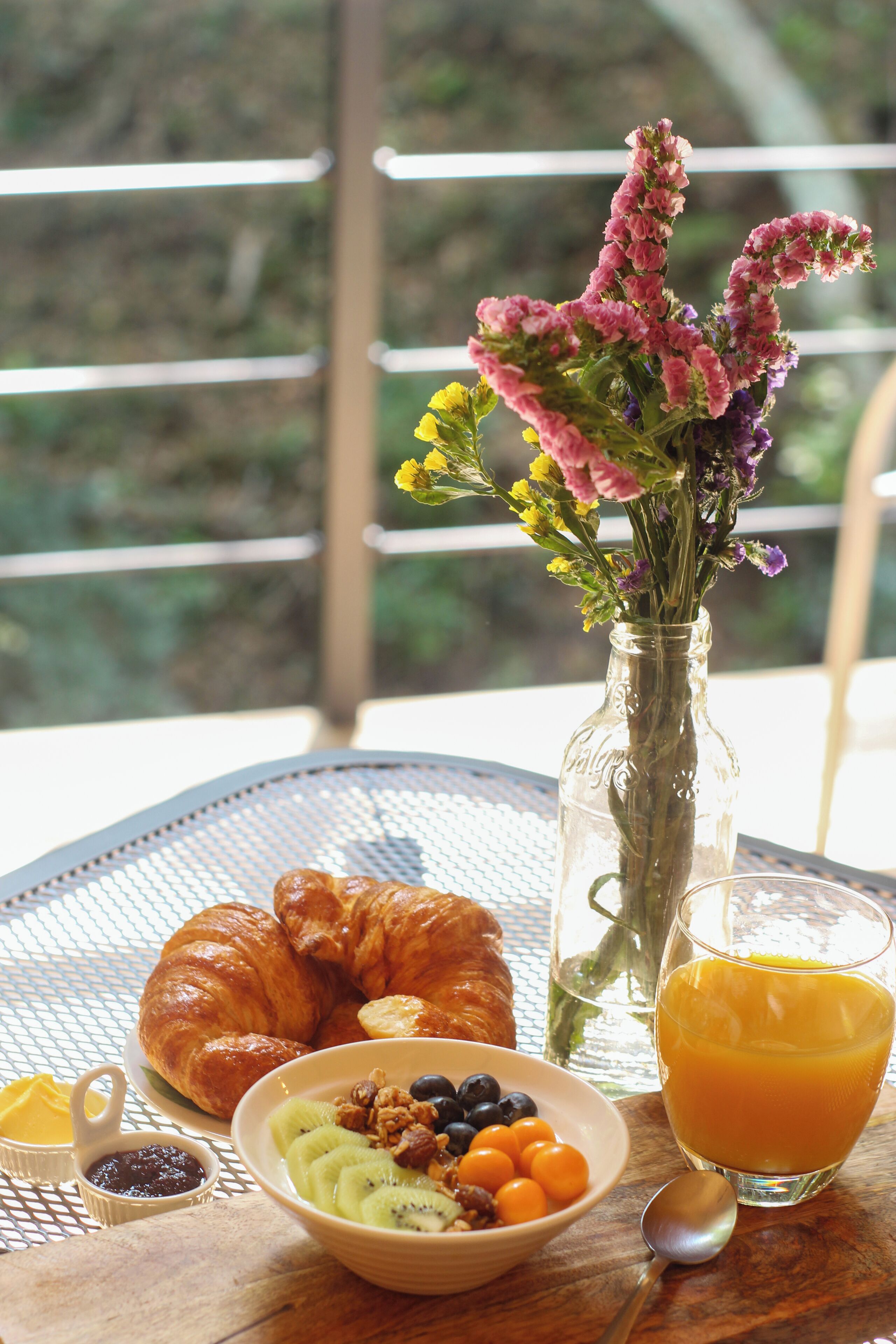 Una mesa de desayuno con croissants, un bol de fruta, un vaso de zumo de naranja y un jarrón de flores.