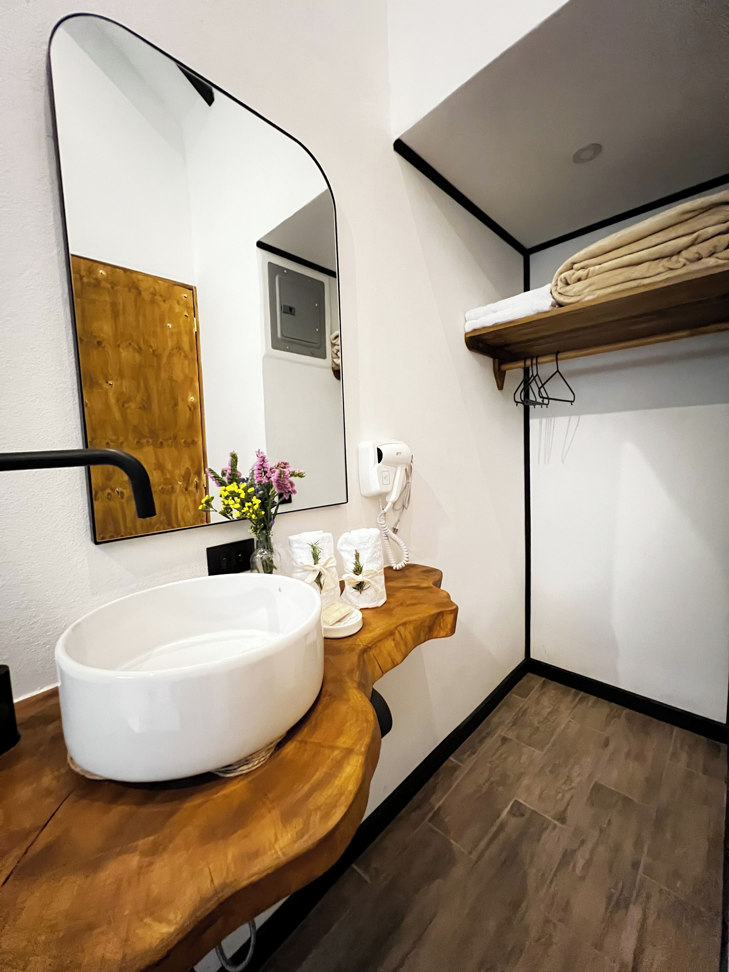 Un baño con lavamanos blanco, tocador de madera, un espejo grande, un perchero para toallas y suelo de madera.