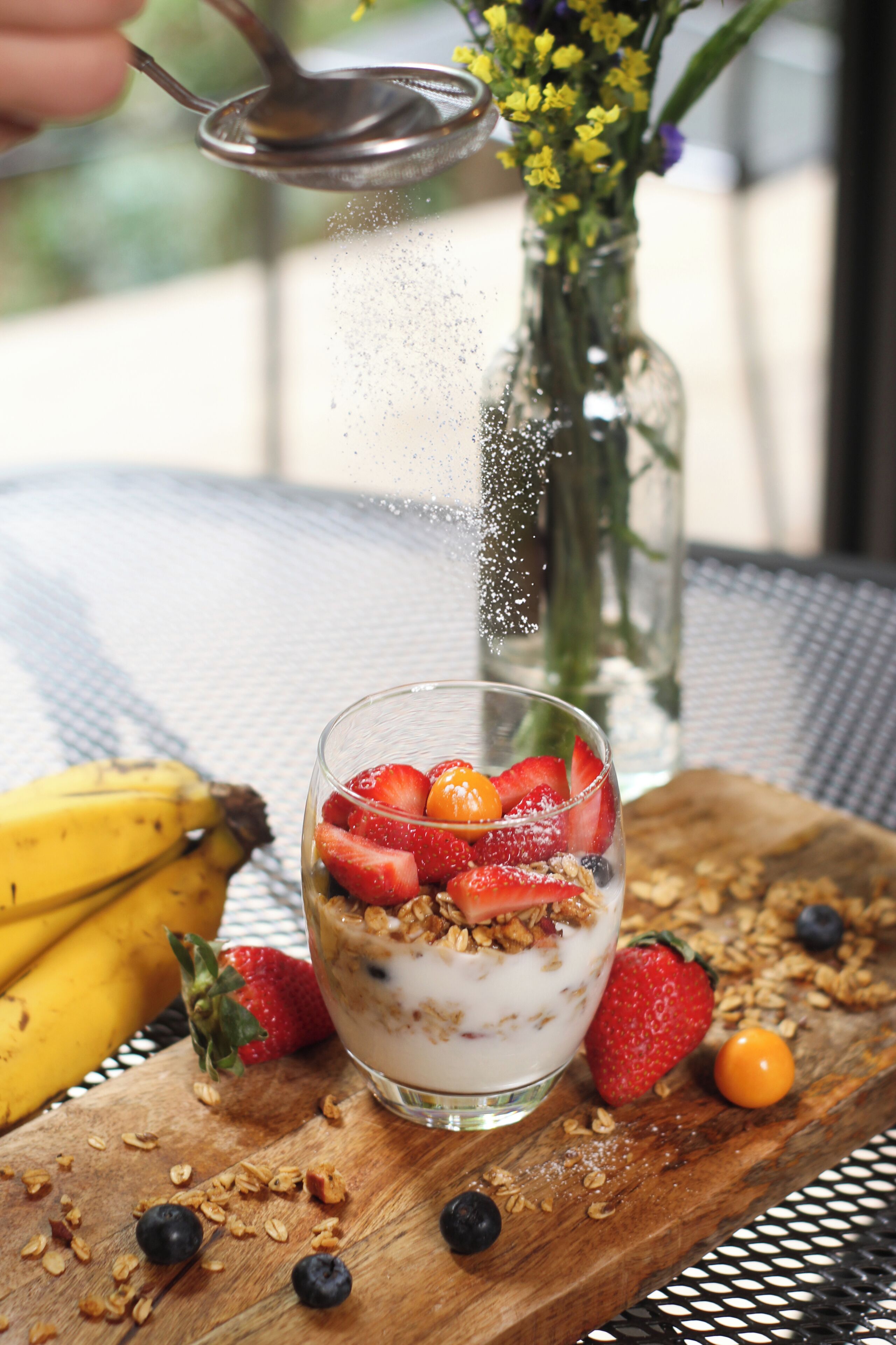 Un vaso de yogur con fresas y granola, un plátano y una cereza sobre una tabla de madera.