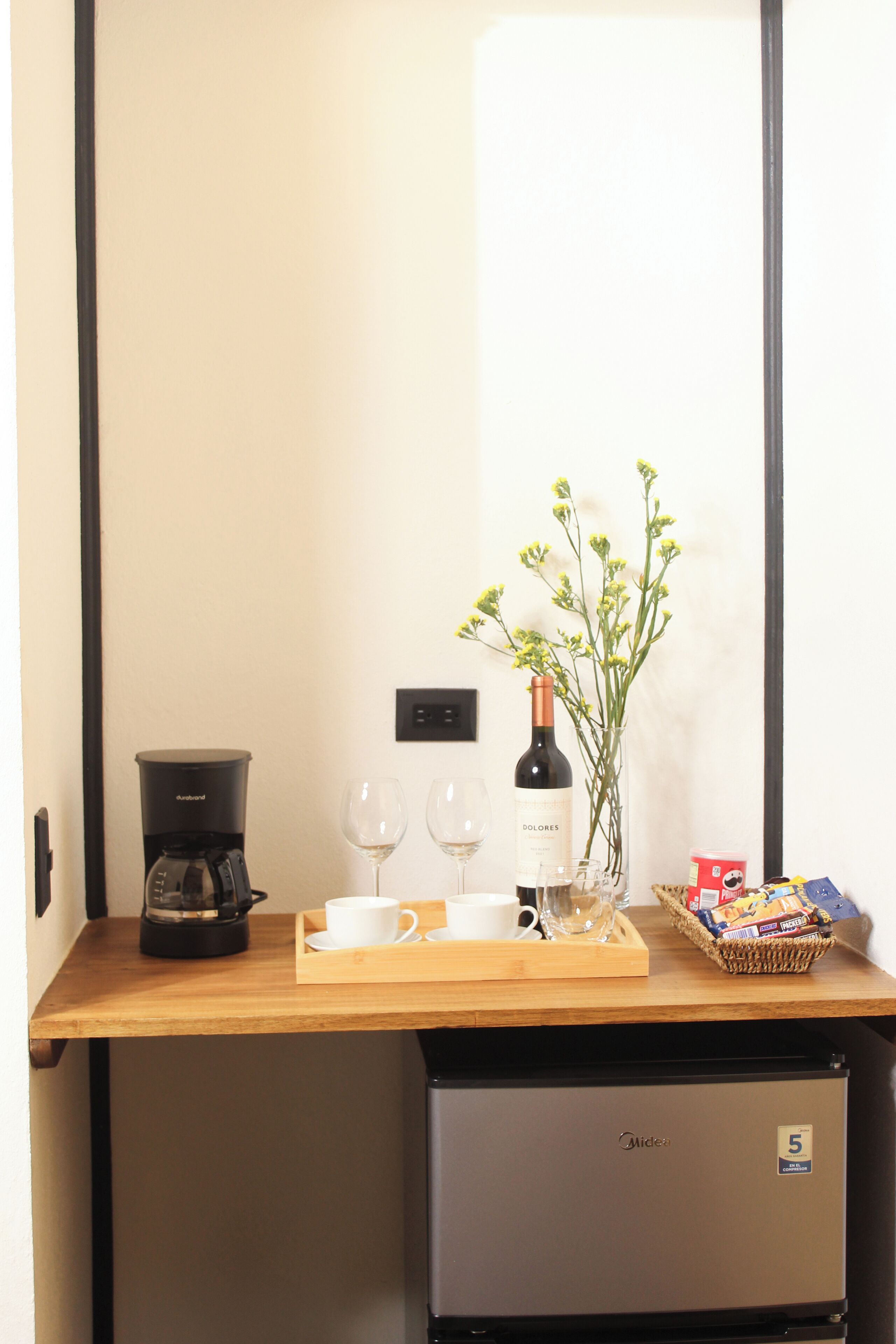 A compact kitchenette with a coffee maker, wine bottle, glasses, and a basket of snacks.