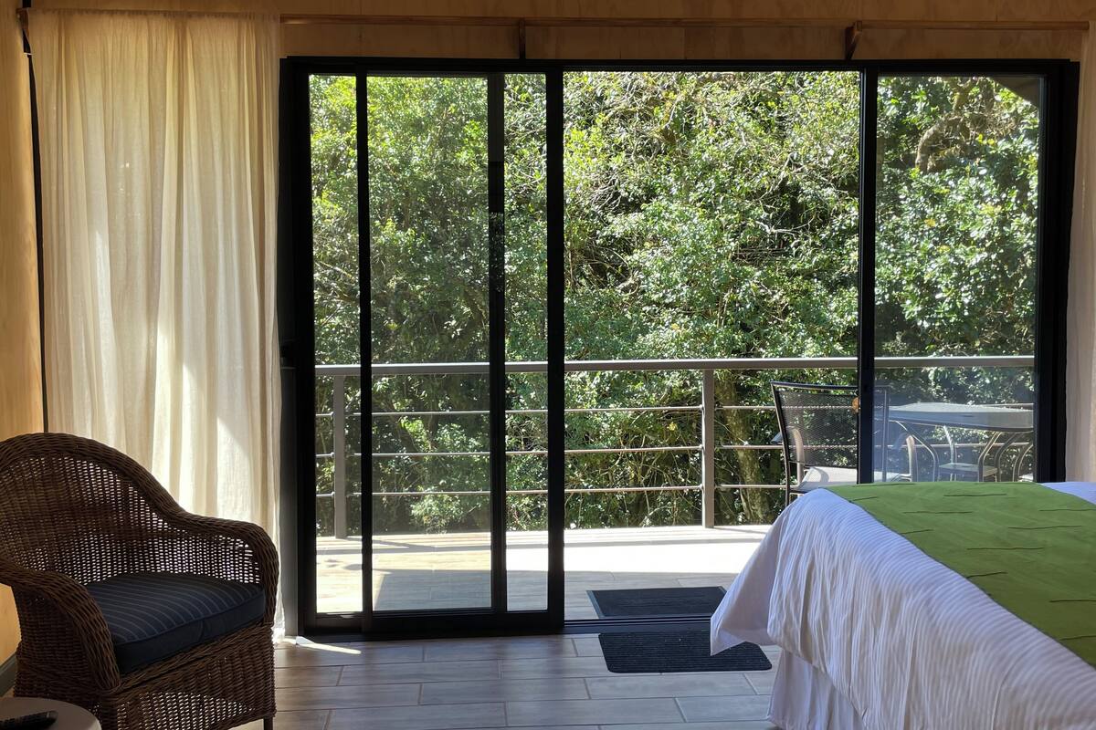 Ein Zimmer mit einem Bett, einem Korbstuhl und einem Balkon mit Blick auf Bäume.