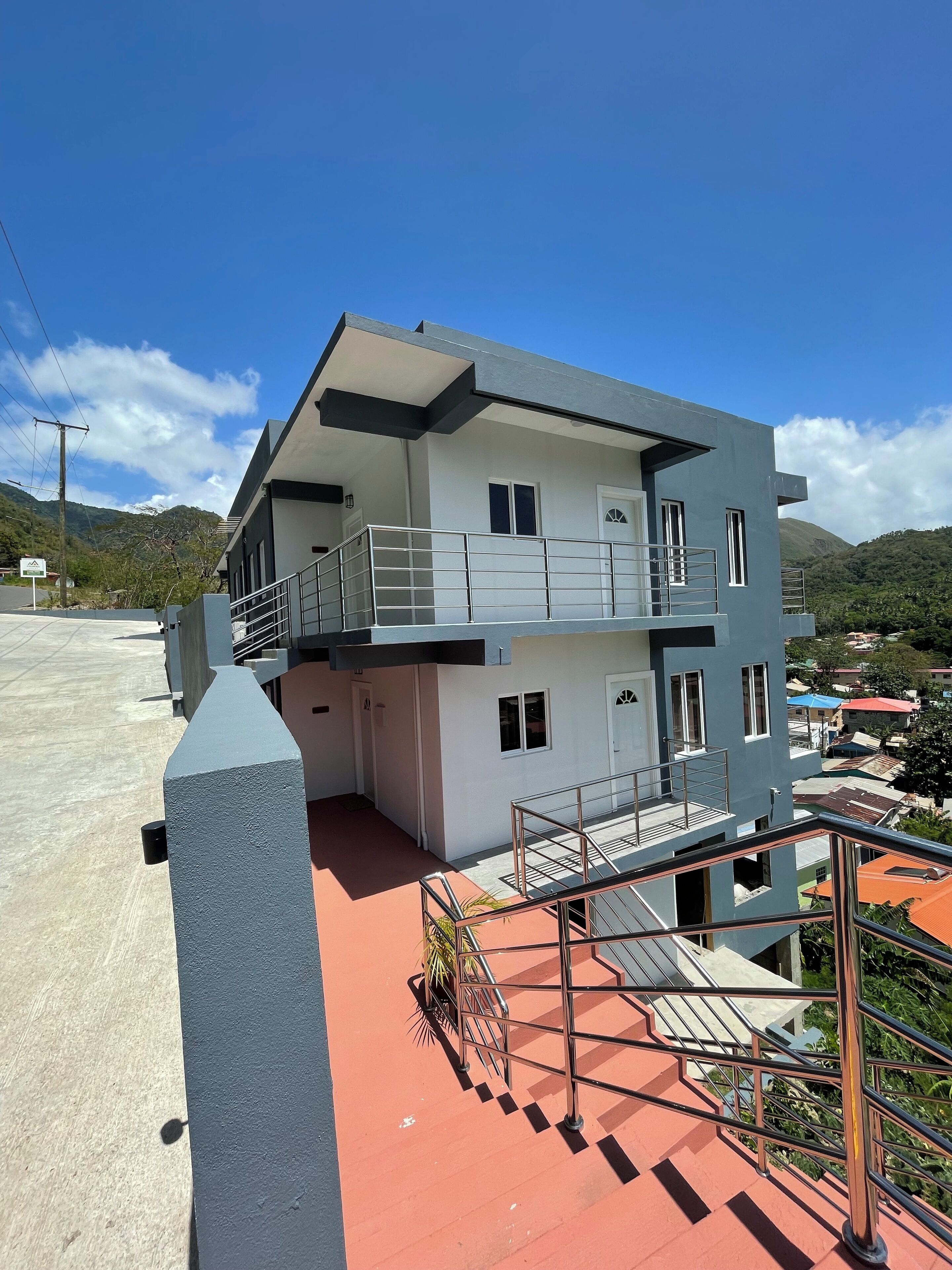 Un edificio moderno de dos pisos con balcón, entrada de tejas rojas y vista de casas y colinas circundantes.