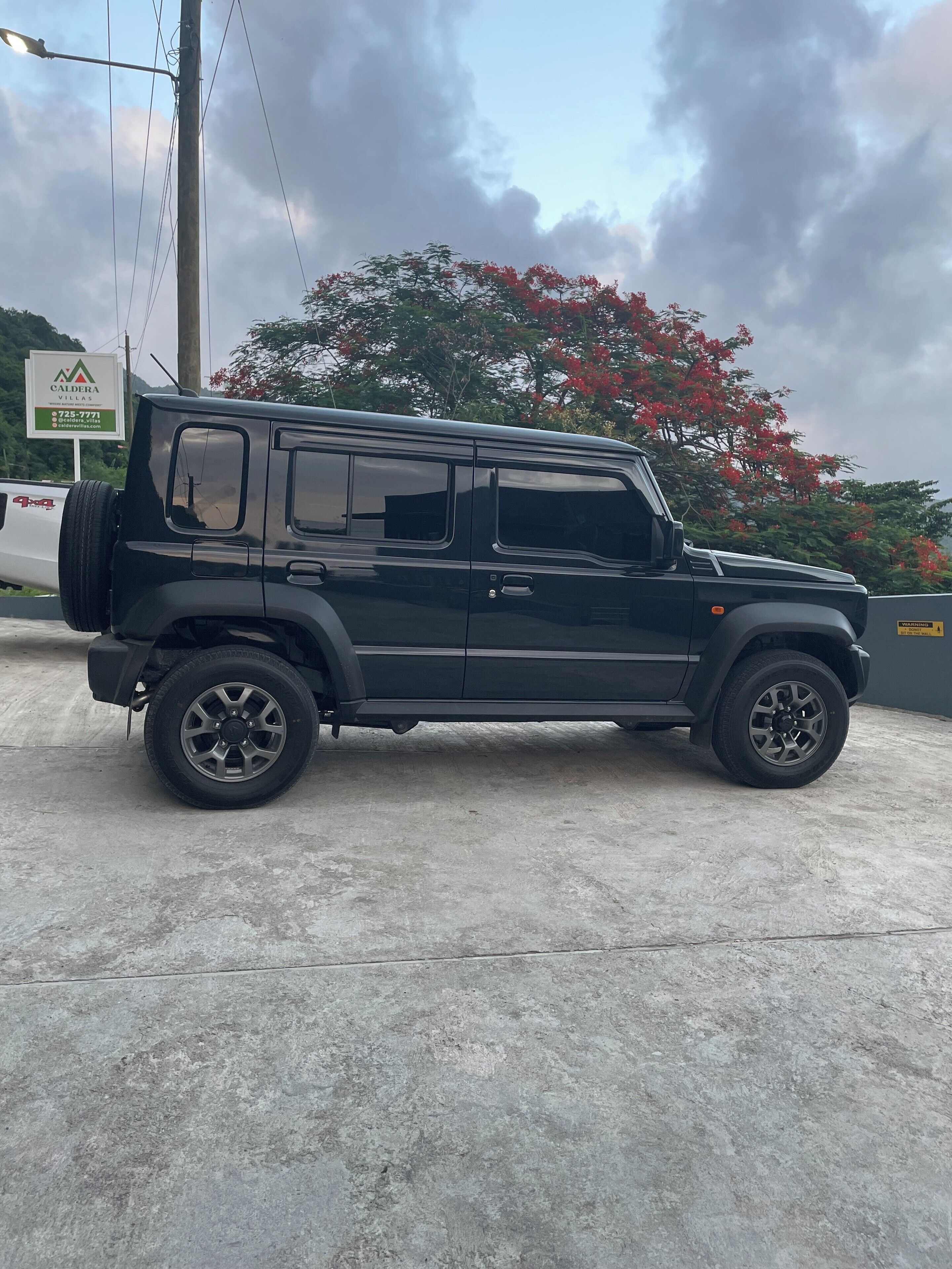 Un SUV negro estacionado en un estacionamiento de piso de concreto, un letrero de Caldera Villas, y un árbol con flores rojas al fondo.