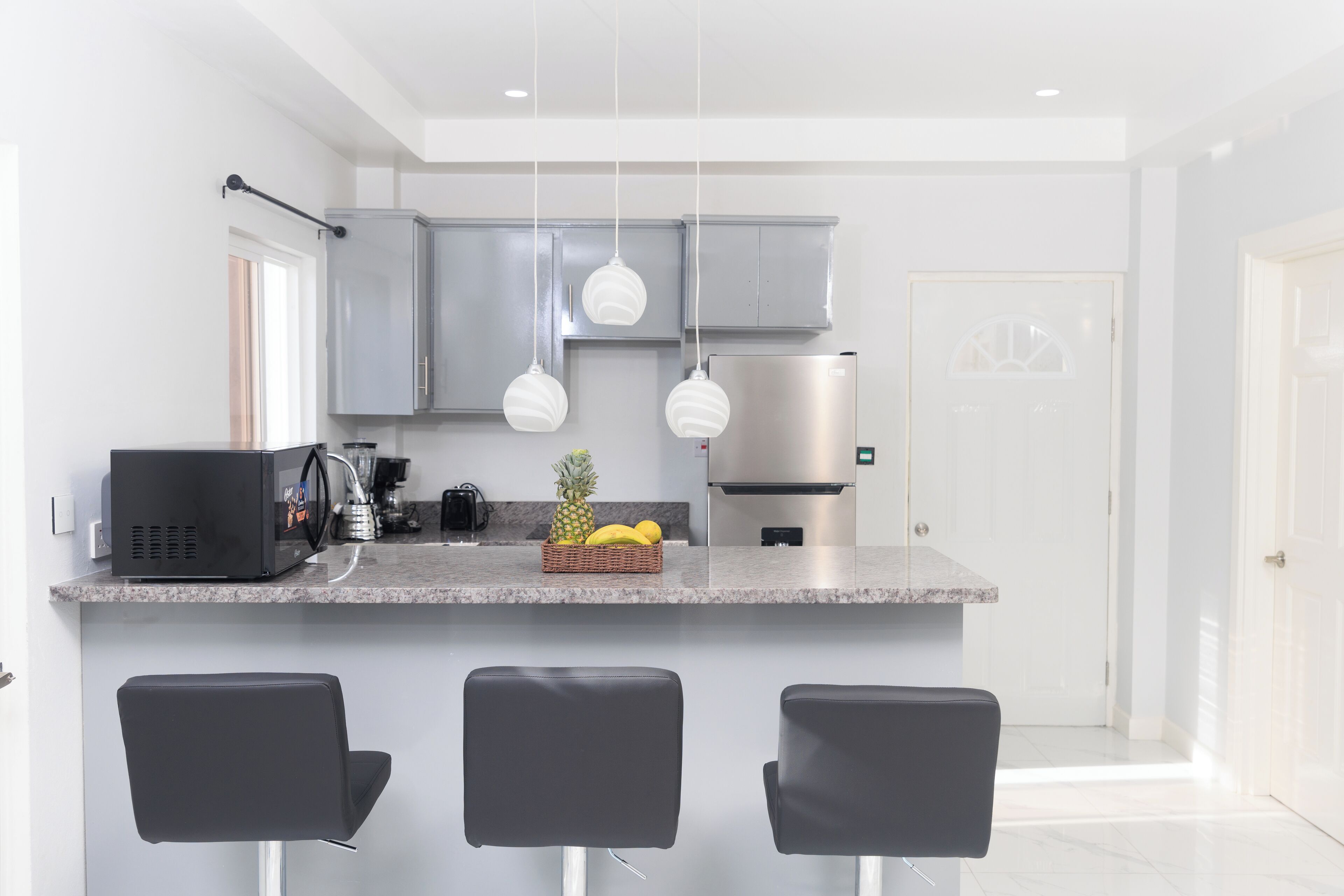 A modern kitchen with grey cabinets, a stainless steel refrigerator, a coffee machine, and a fruit basket on the counter. There are three black bar stools in front of the counter.
