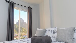 A bedroom with a bed, curtains, and a view of a mountain and buildings. - Caldera Villas (Soufrière)